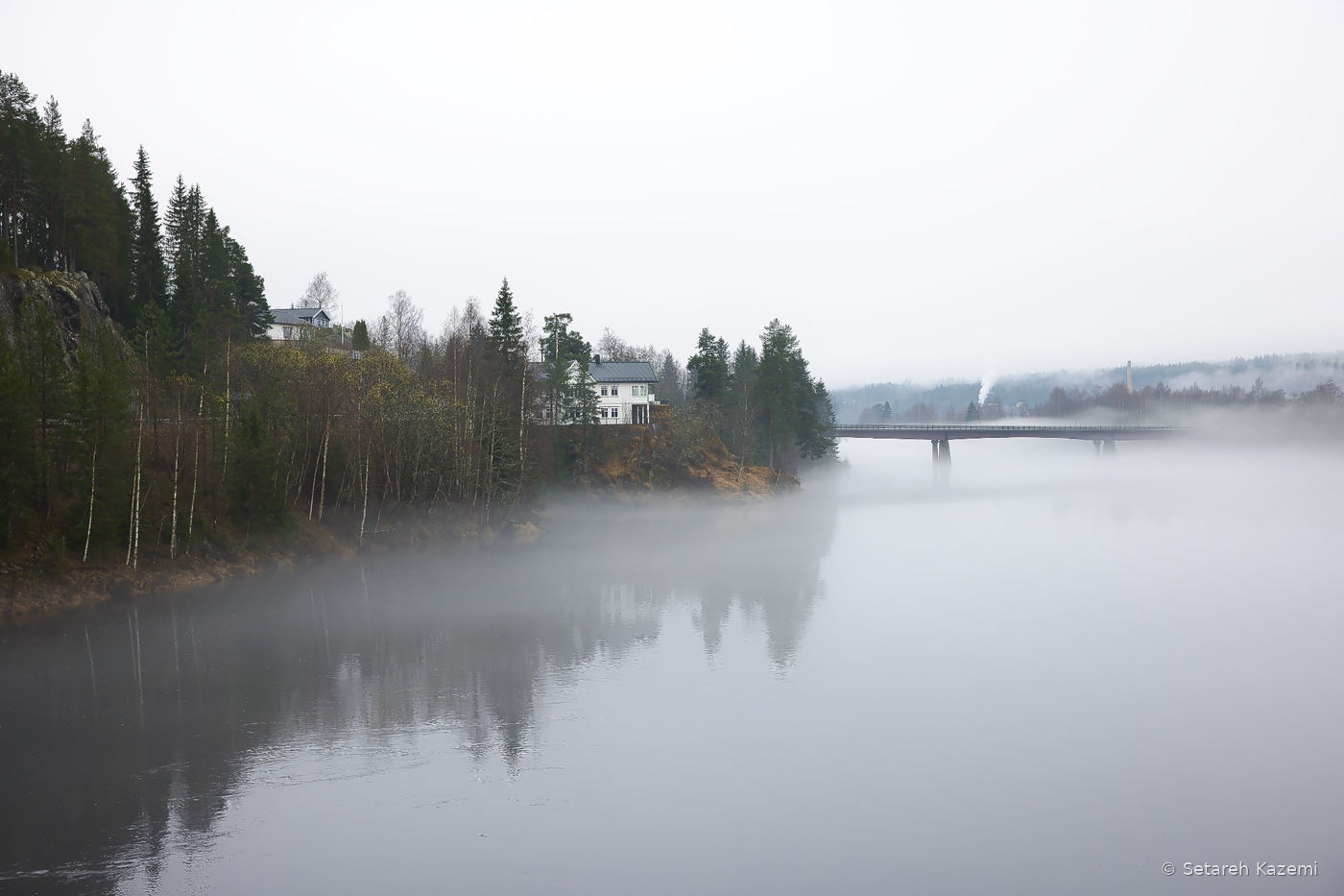 Misty river landscape with a bridge, forest and white houses.